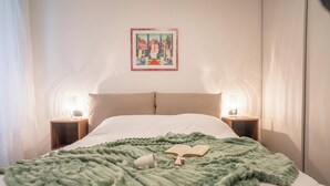 A neatly made bed with a green blanket, a book, and a cup on it. There are two bedside tables with lamps and a framed picture on the wall above the bed. - Pendulum - 9 (Siena)