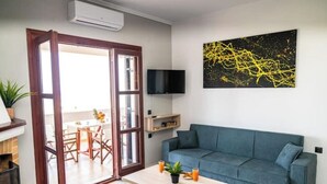Ein Wohnzimmer mit einem blauen Sofa, einem Holztisch mit zwei Gläsern und einer Pflanze, einem Fernseher an der Wand und einem großen, abstrakten Gemälde.