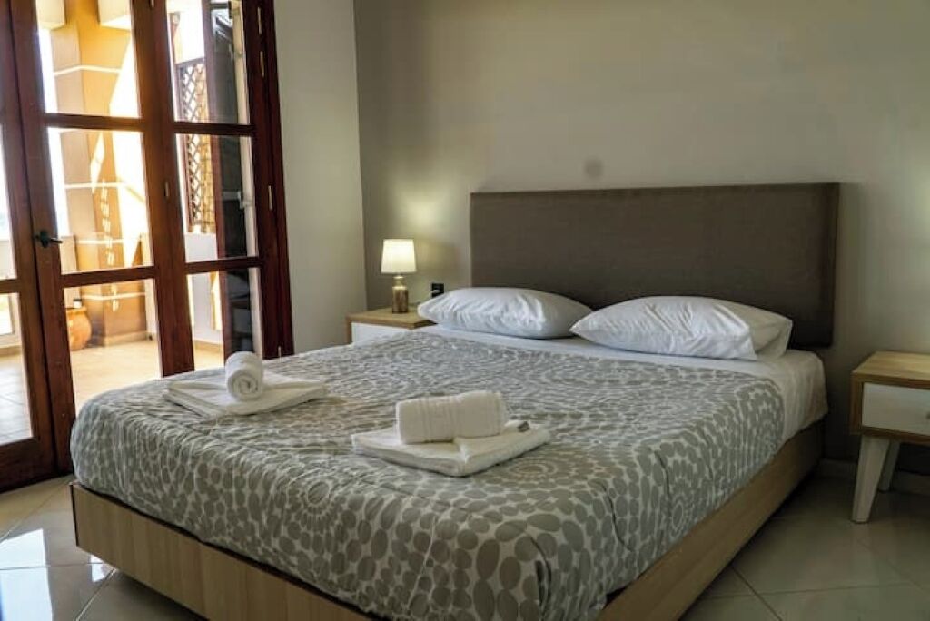 Habitación de hotel con cama, mesita de noche, lámpara y vista a través de una puerta de cristal.