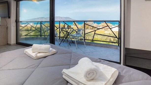 A modern hotel room with a large bed, a balcony with a view of the sea, and a dining area with chairs. - Relax in Style at Ben’s Homes Panorama (Nopigia)