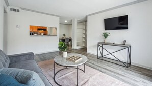 Ein modernes Wohnzimmer mit einem grauen Sofa, einem runden Couchtisch mit Pflanze, einem an der Wand montierten Fernseher und einem Essbereich im Hintergrund.