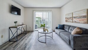 A modern living room with a sofa, a coffee table, a TV mounted on the wall, and a large window with blinds.