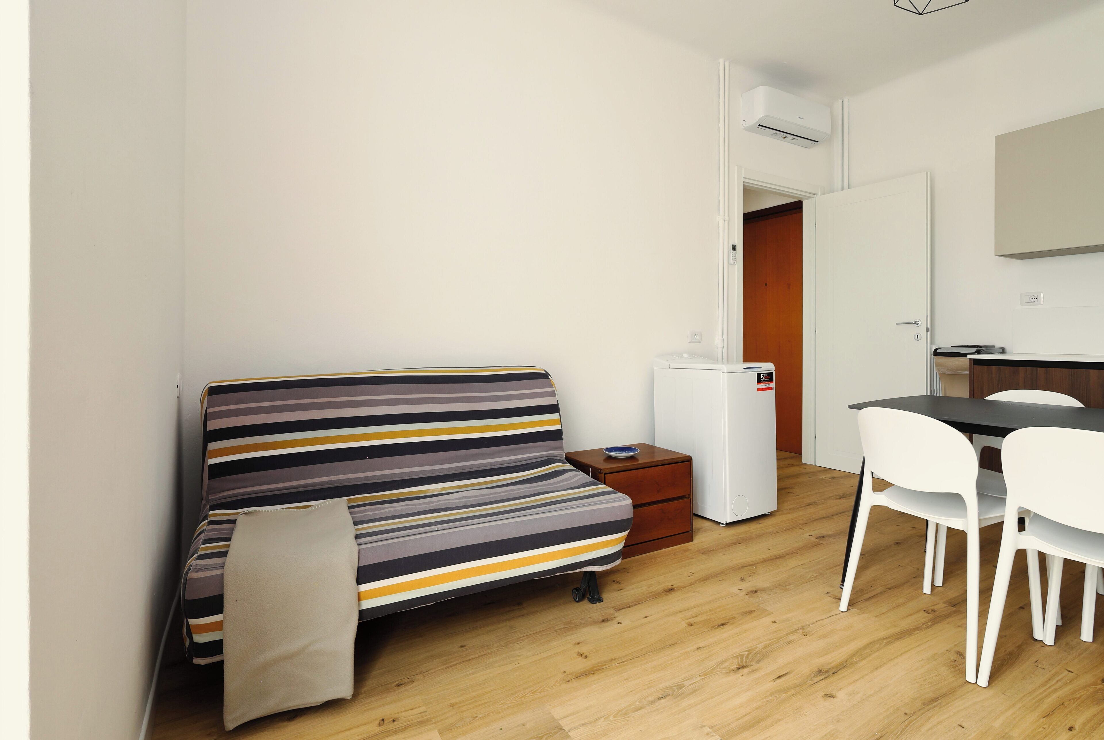 A small, modern room with a striped sofa, a compact dining table with chairs, a small refrigerator, and a door leading to another room.
