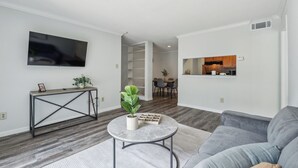 Un salon moderne avec un canapé gris, une table basse ronde agrémentée d’une plante, un téléviseur fixé au mur et un coin cuisine visible à travers une porte ouverte.