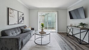 Un salon moderne avec un canapé gris, une table basse ronde agrémentée d’une plante, un téléviseur fixé au mur et une porte-fenêtre coulissante donnant sur un balcon.
