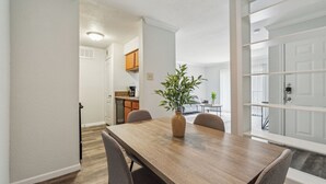 Un coin repas avec une table et des chaises en bois, un vase avec une plante, et une vue sur la cuisine et le salon par une porte ouverte.