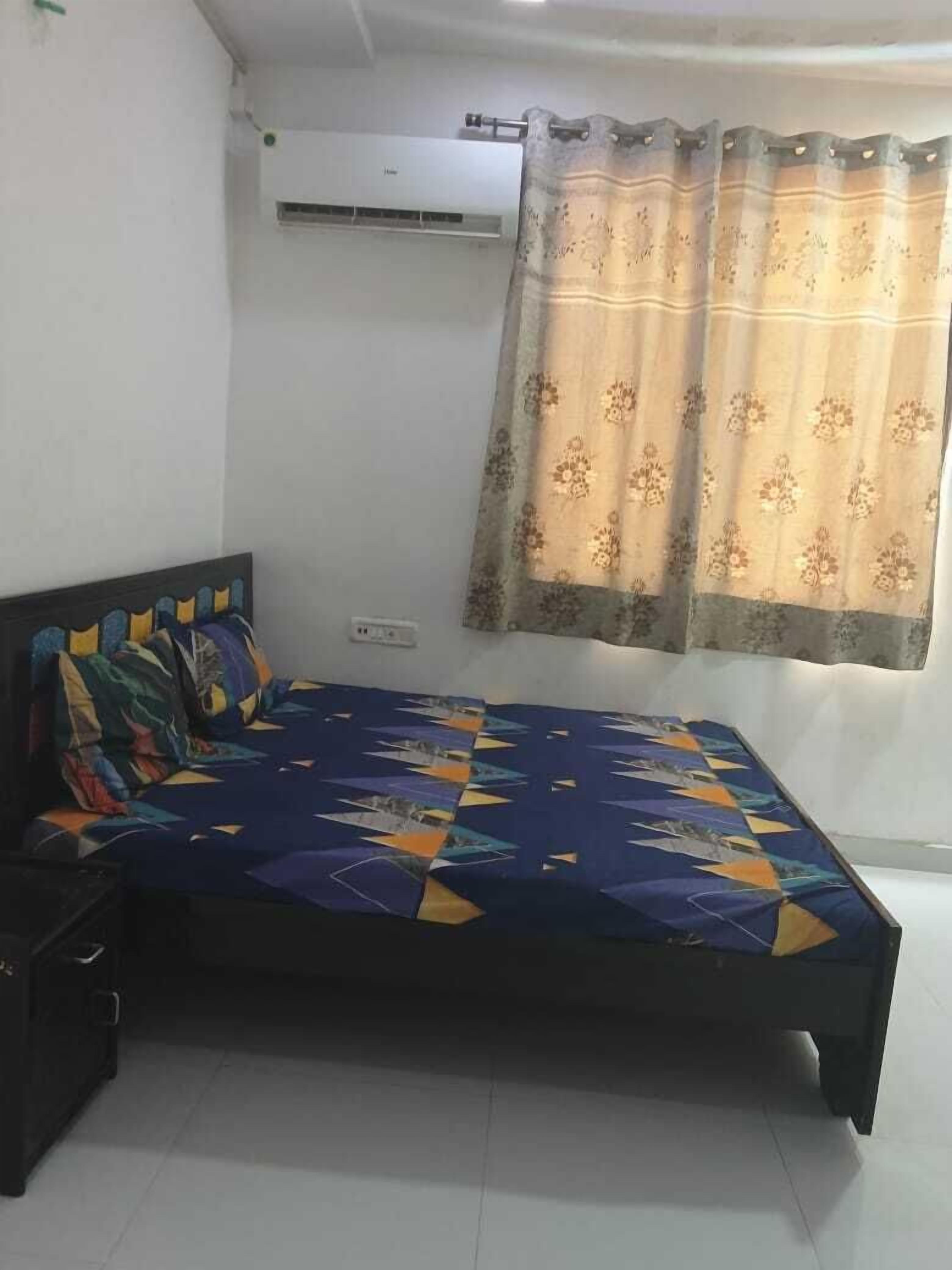 A bedroom with a bed, a nightstand, a wall-mounted air conditioner, and a window with a patterned curtain.