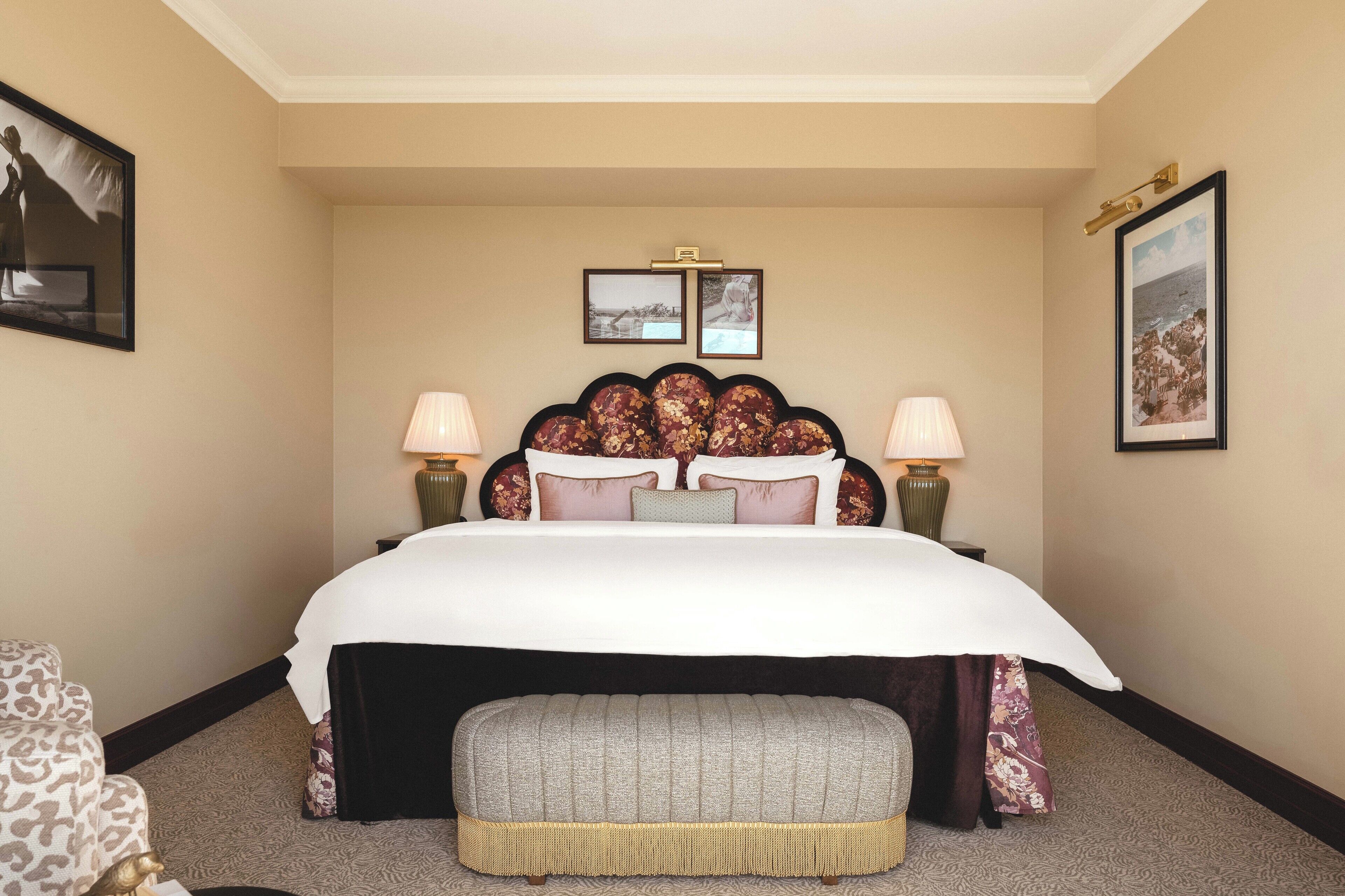 Una habitació d'hotel amb un llit gran, dues làmpades de nit, un capçal de llit amb decoracions florals, una petita banca i quadres a les parets.