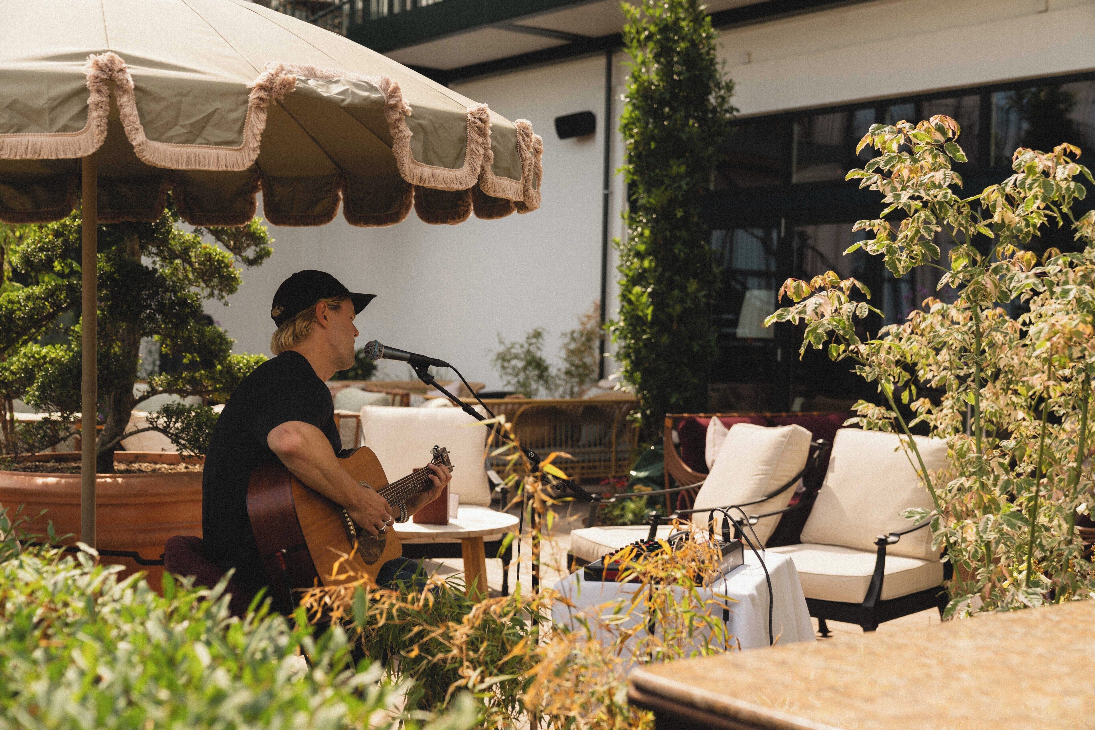En person spiller guitar på en udendørs café med borde og stole, omgivet af planter og en parasol.