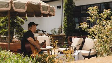 En person spiller guitar på en udendørs café med borde og stole, omgivet af planter og en parasol.