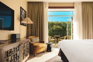 A hotel room with a bed, a TV, a desk, a chair, a lamp, and a balcony with chairs.