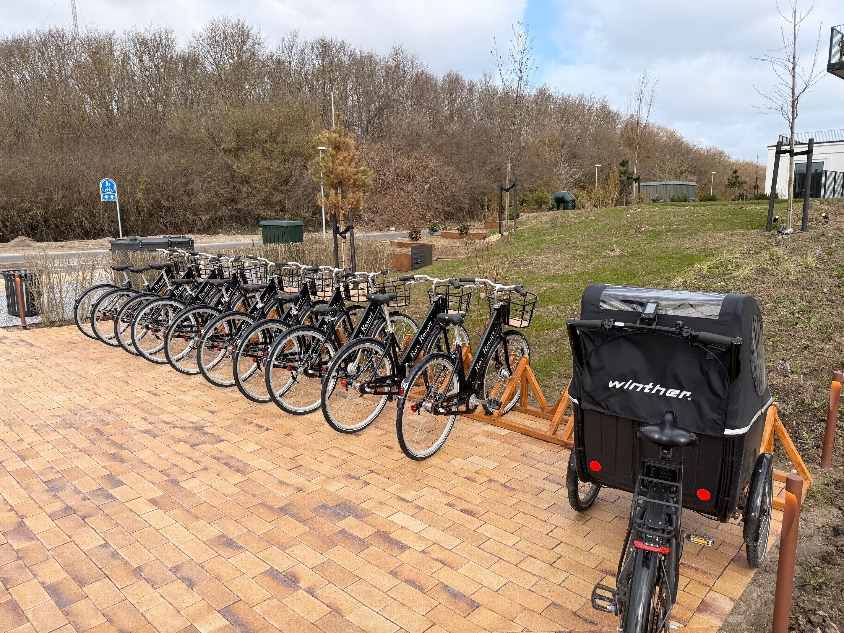 Varias bicicletas aparcadas sobre una superficie de ladrillo, un aparcamiento cubierto para bicicletas y un pequeño carro con una cubierta negra.
