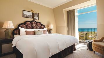 A hotel room with a large bed, two bedside tables with lamps, a view of the sea, and a balcony with outdoor furniture.