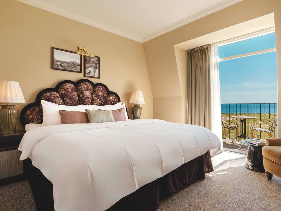 A hotel room with a large bed, two bedside tables with lamps, a view of the sea, and a balcony with outdoor furniture.