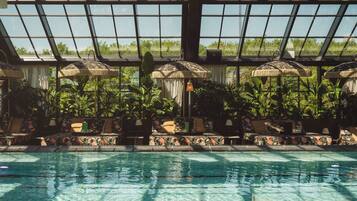 An indoor swimming pool with lounge chairs and umbrellas, surrounded by lush greenery and a glass ceiling.