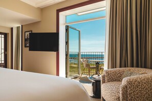 A hotel room with a bed, a chair, a small table, a TV, and a balcony with a view of the sea.