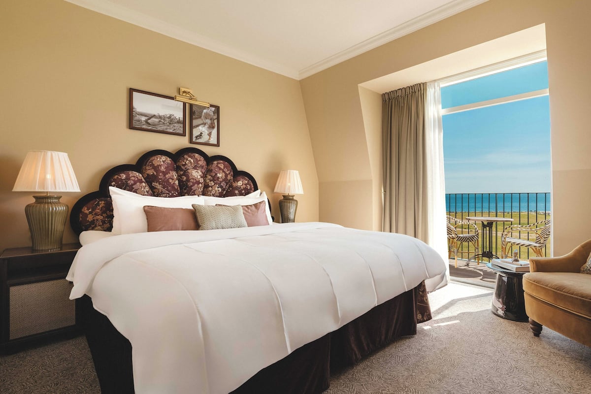 Habitación de hotel con una cama grande, dos mesitas de noche con lámparas, vista al mar y un balcón con mobiliario exterior.
