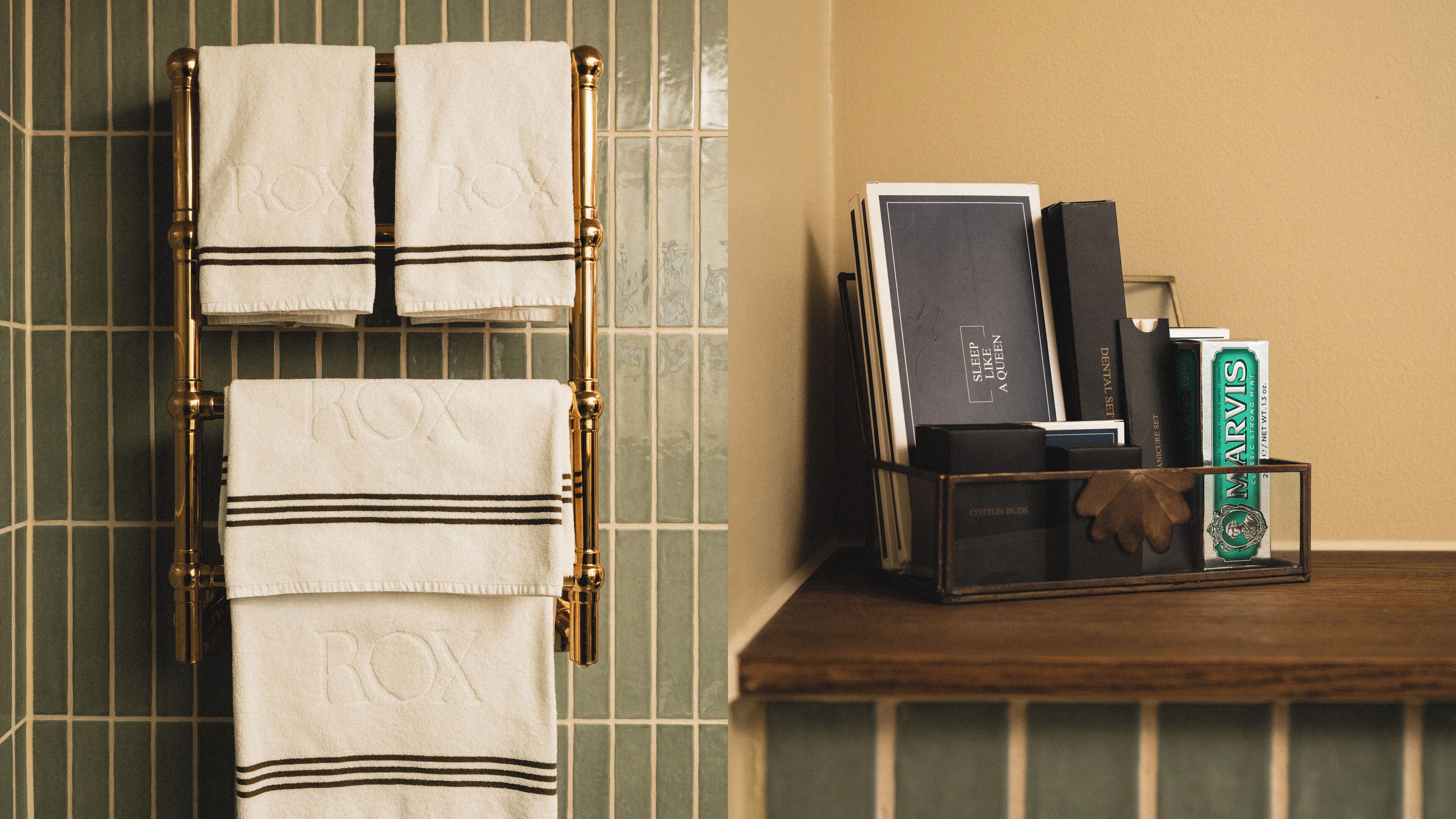 A bathroom with a towel rack holding two towels, a shelf with books and a box of tea, and a tiled wall.