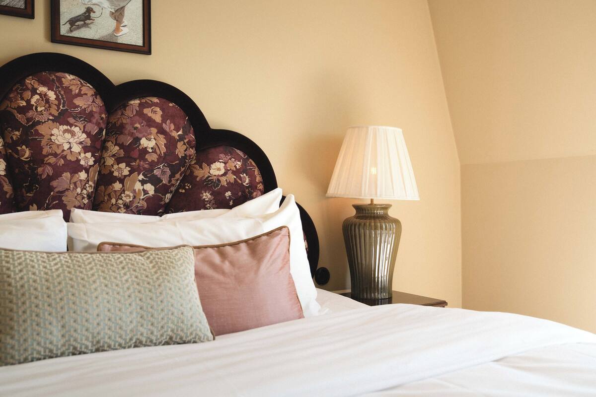 Una cama bien hecha con almohadas estampadas con motivos florales y una lámpara en una mesita de noche.