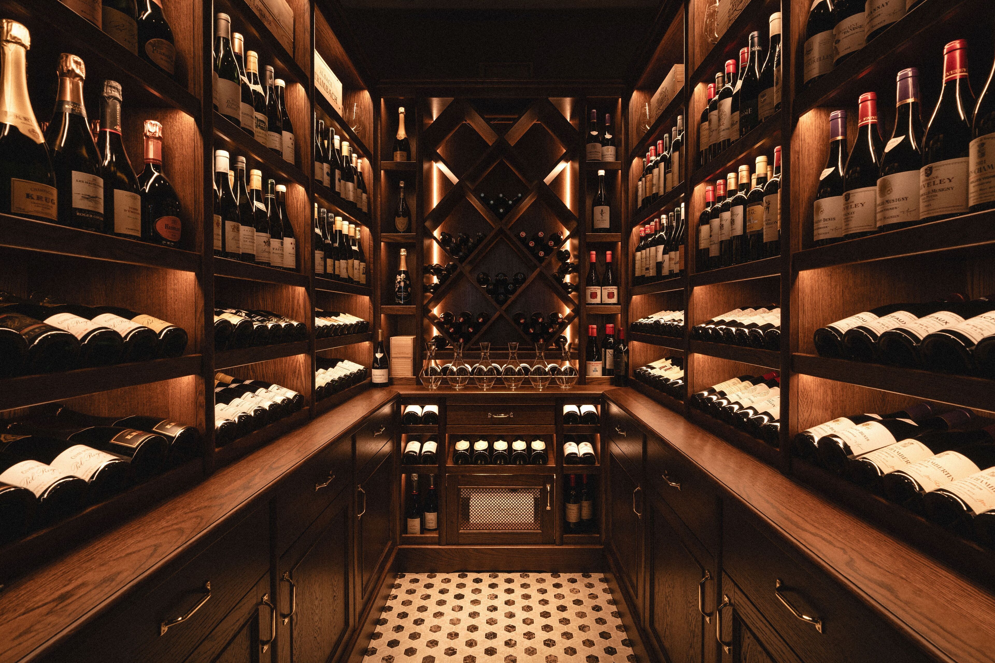Una bodega con estanterías y botelleros de madera, que alberga numerosas botellas de vino y copas.