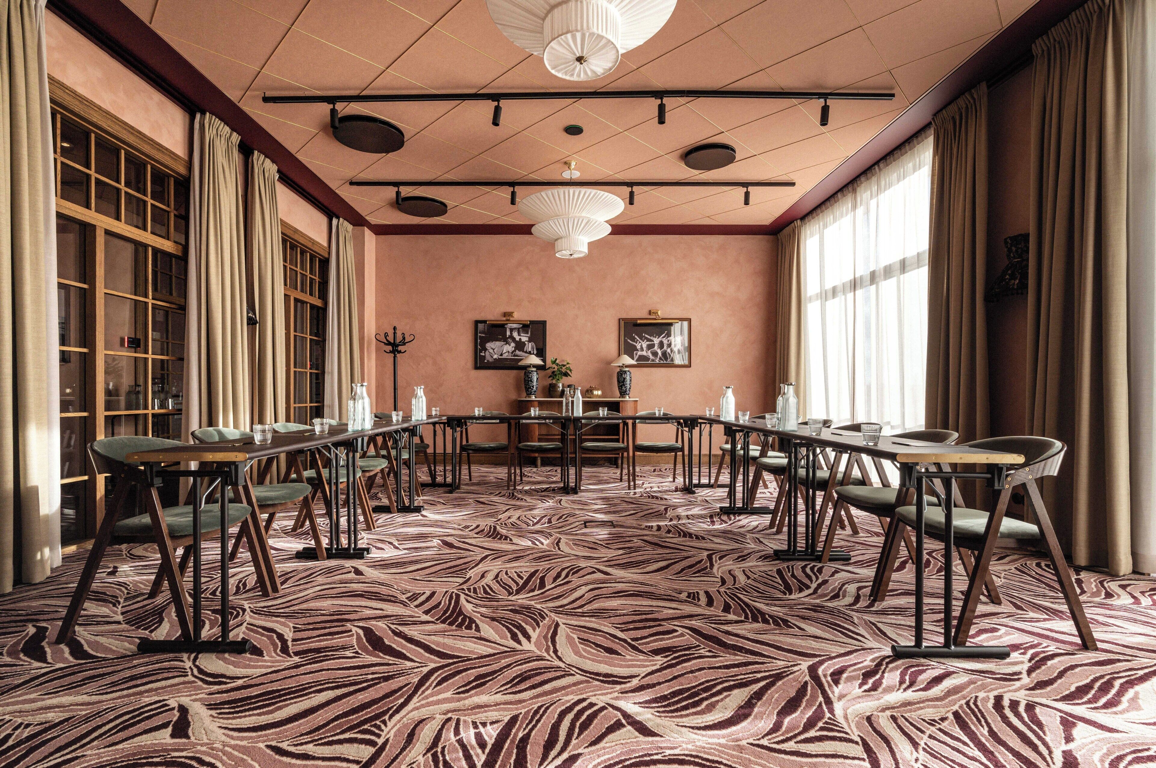 Ein Konferenzraum mit Teppich in Zebramuster, Holztischen und -stühlen, großen Fenstern mit Vorhängen und einer Kristallleuchter.