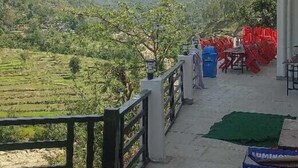 A balcony with a view of green fields and mountains. There are tables and chairs outside. - Hotel Riyansh  (Ukhimath)