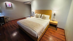 Habitación de hotel con una cama grande, suelo de madera, cabecera, lámparas de noche y una mesita.
