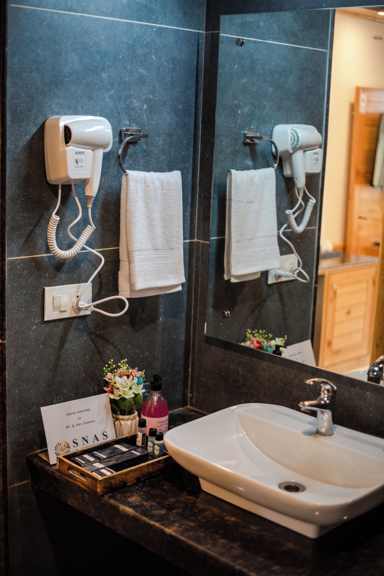 A bathroom with a sink, two hair dryers mounted on the wall, a towel rack, and a welcome sign.