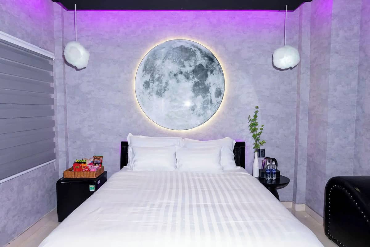 Une chambre avec un lit, un luminaire rond en forme de lune fixé au mur, une petite table d’appoint avec une plante et une chaise noire.