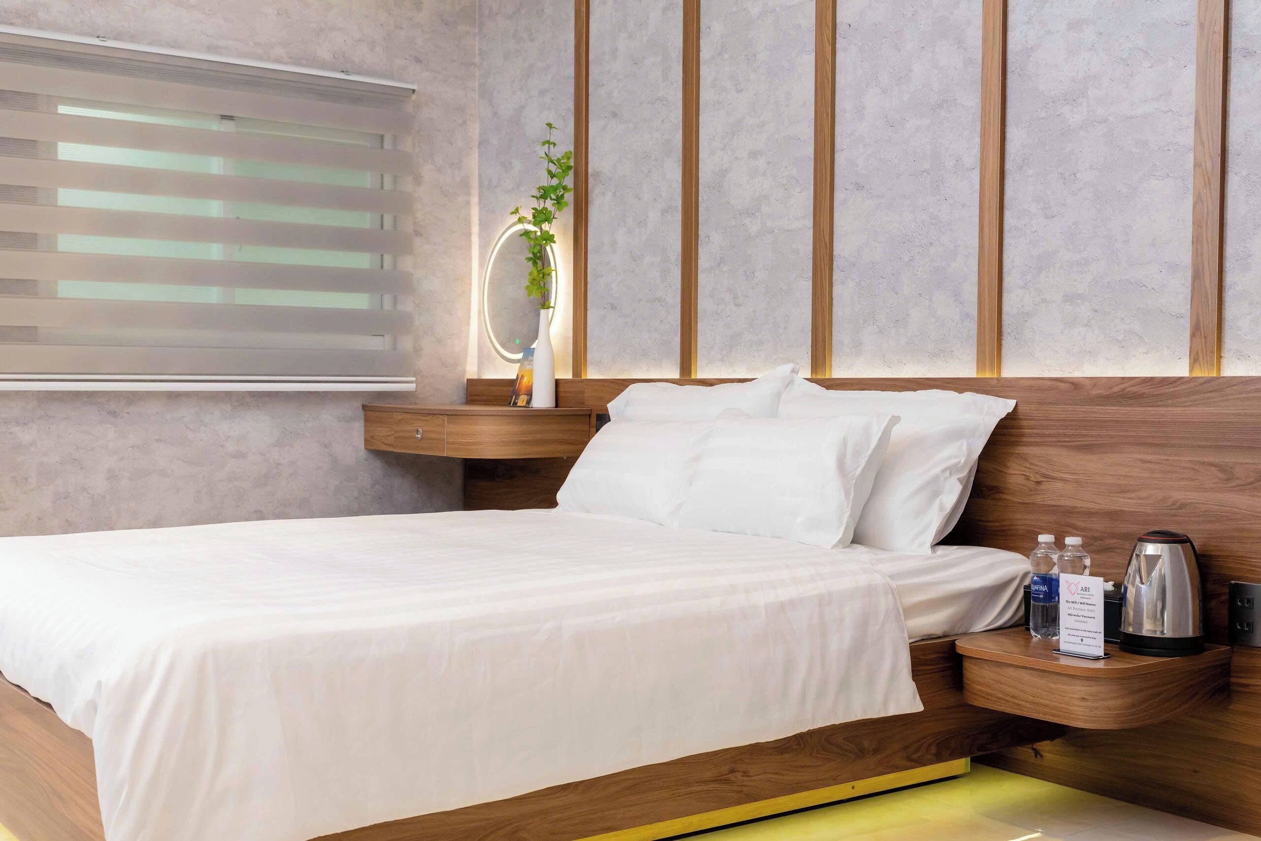 A modern bedroom with a wooden headboard, a bedside table with a mirror, a small plant, and a nightstand with a kettle and water bottles.
