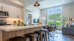 A modern kitchen with a central island, bar stools, a dining area with a view, and a living room with a sofa and lamp. - Apres Ski | DT | MtnView | TopFloor | AC | Sleep 6 (Golden)