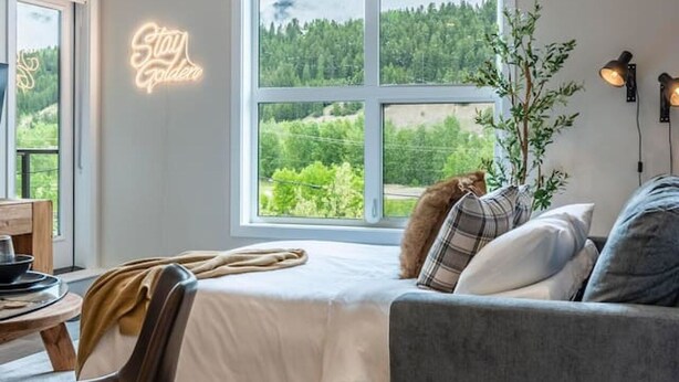 A cozy room with a large window, a bed, a grey sofa, a small table, and a neon sign that reads "Stay Longer". - Apres Ski | DT | MtnView | TopFloor | AC | Sleep 6 (Golden)