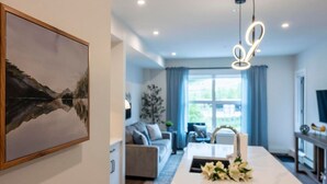 A modern living room with a sofa, a dining area with a table and flowers, and a large window with curtains.