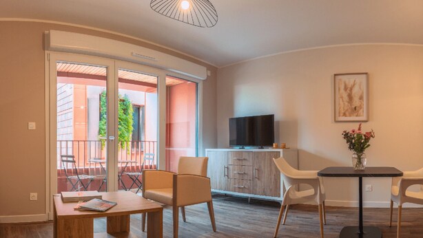 A modern living room with a wooden coffee table, two armchairs, a TV on a wooden cabinet, and a balcony with a view of greenery. - Genius: An exceptional residence in Besançon in the heart of the Viotte business (BESANÇON)