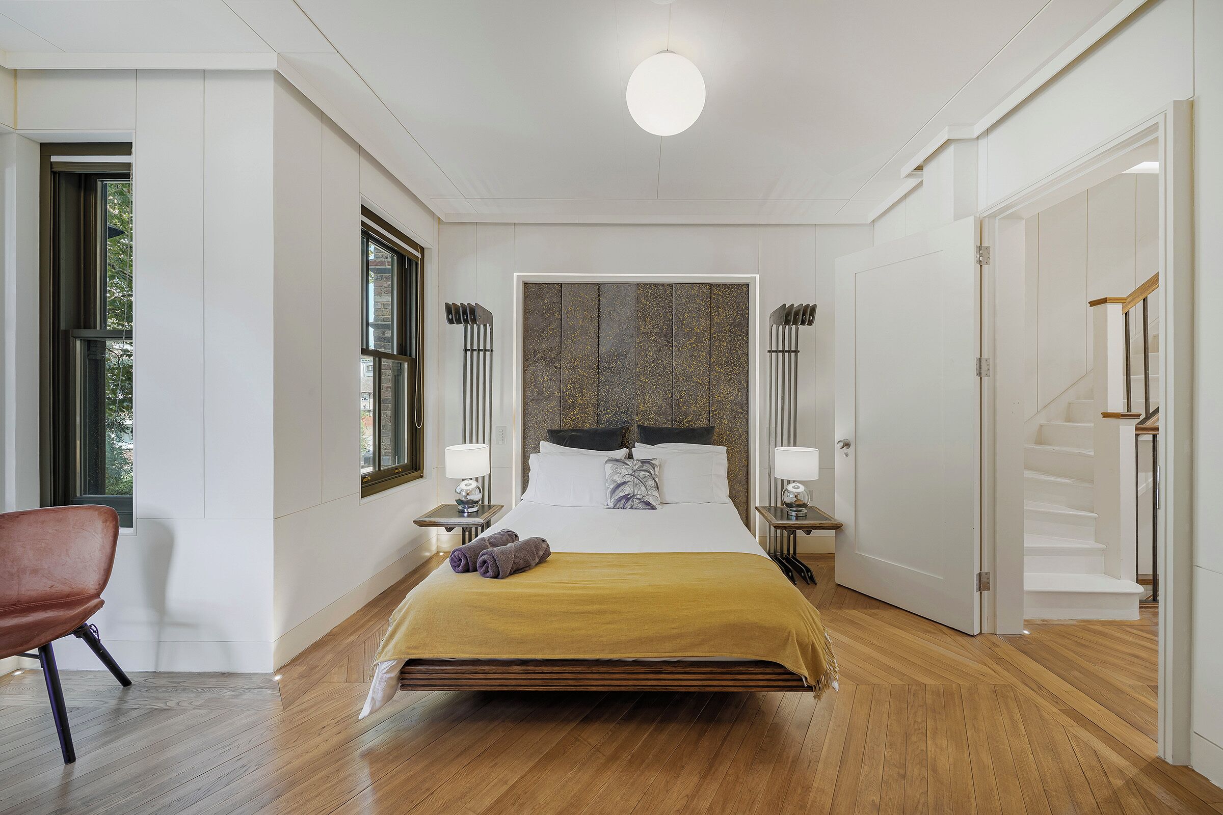 Un dormitorio moderno con una cama grande, piso de madera, una silla roja y vistas al exterior a través de una ventana.