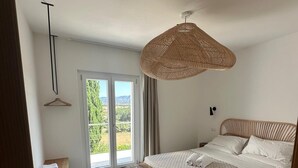 A modern bedroom with a large bed, a hanging rattan light fixture, a bench, and a view of greenery through the sliding glass door. - Agreen Country House Myrtle (Sardegna)