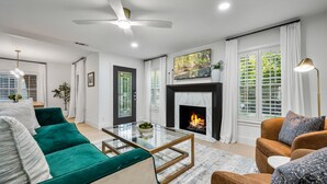 A modern living room with a fireplace, green and brown sofas, a glass coffee table, and a ceiling fan. - The Lyndzee-Luxury Downtown Haven, Pool, Sleeps 16 (Dallas)
