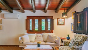 Un salon chaleureux avec un plafond en bois, un canapé beige agrémenté de coussins décoratifs, un fauteuil à motifs floraux, une table basse en bois et une petite table d’appoint avec un vase.