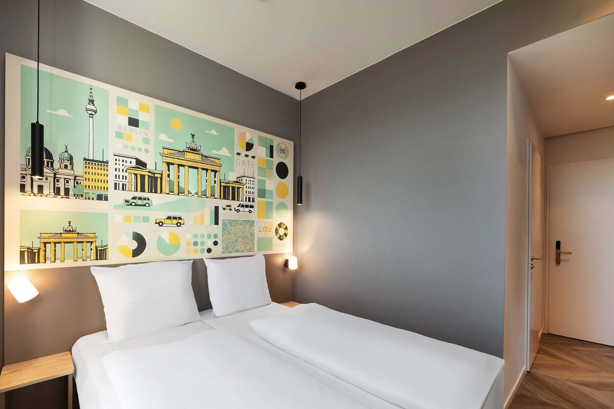 Une chambre d’hôtel moderne avec un grand lit, une table de chevet et une fresque murale représentant des monuments emblématiques de la ville.