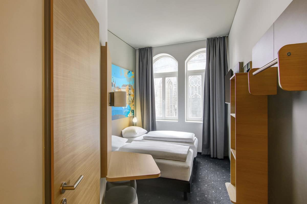 A hotel room with a bed, a desk, a chair, a wardrobe, and a painting on the wall.