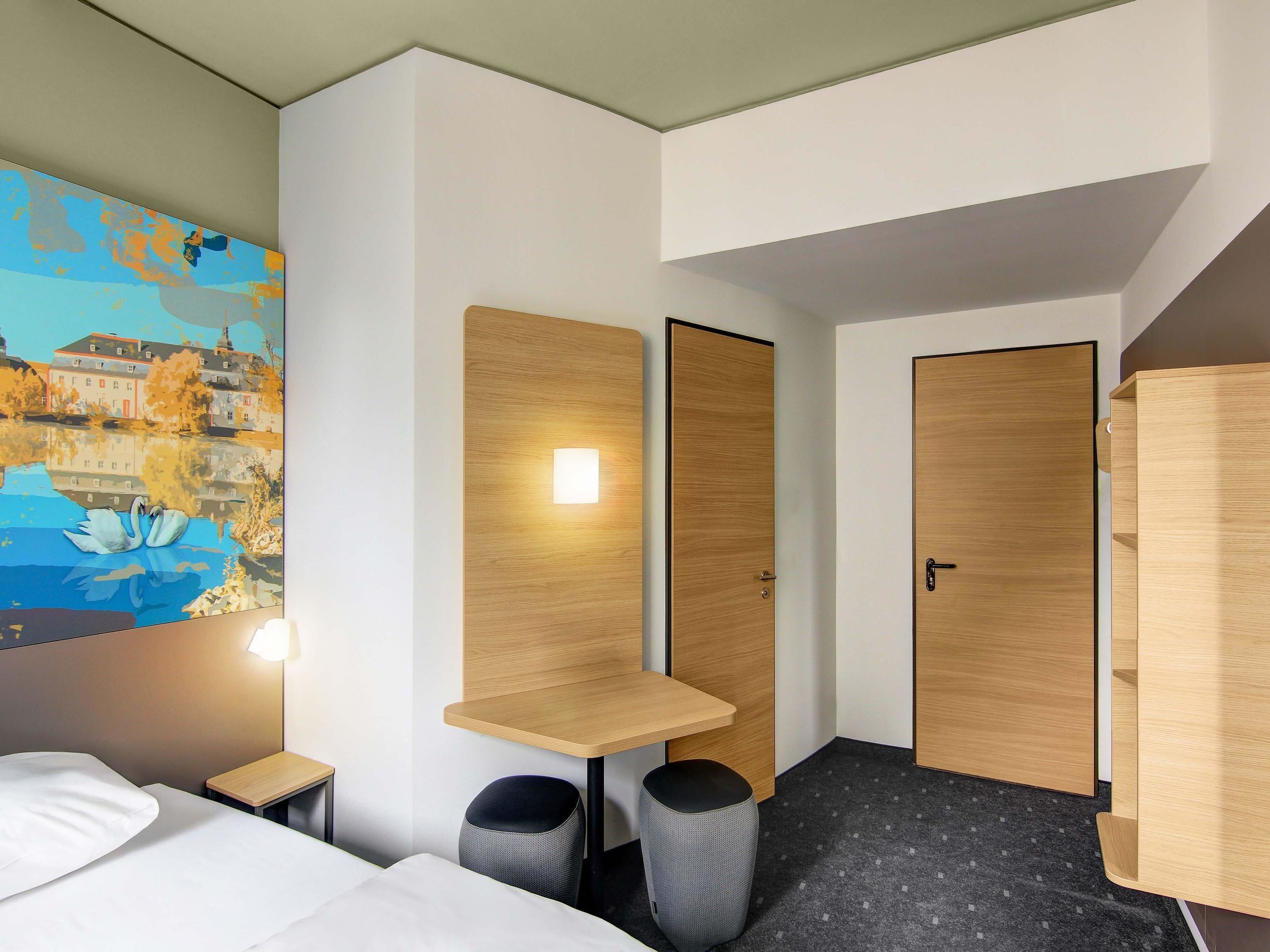 Une chambre d’hôtel moderne équipée d’un lit, d’un bureau, de deux chaises, d’une poubelle et d’un tableau représentant des cygnes sur un mur.
