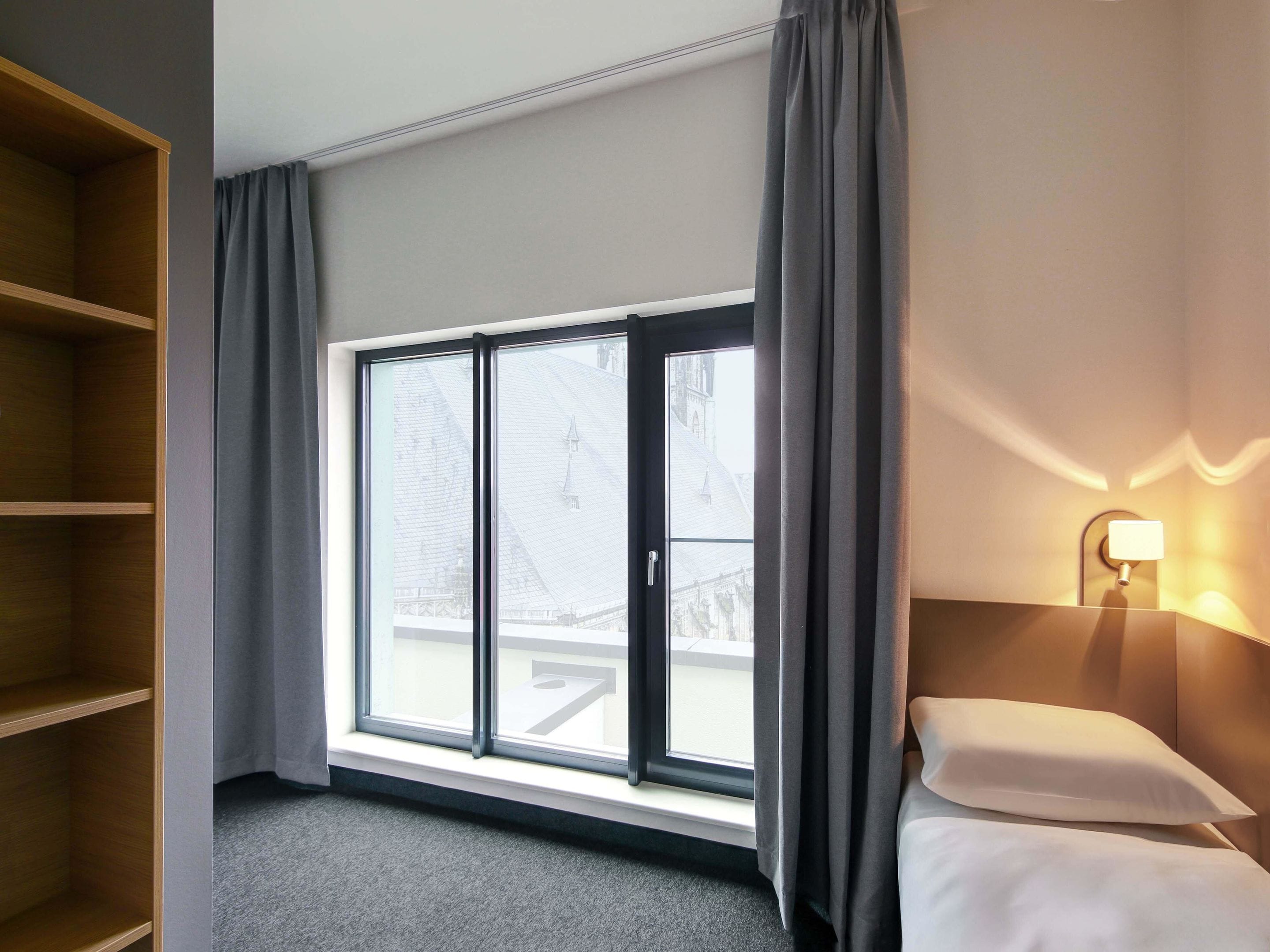A hotel room with a large window, a bed with white linens, a nightstand with a lamp, and a wooden shelf unit.