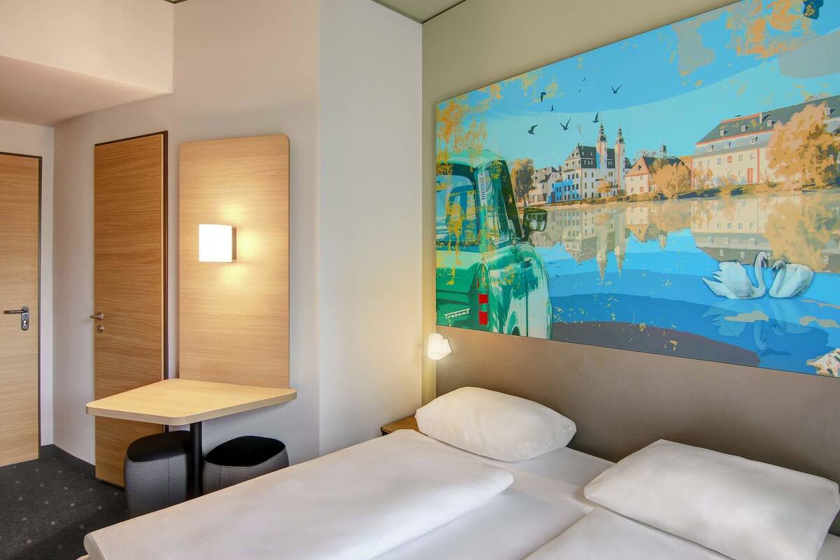 A hotel room with a bed, a small table, and a wall-mounted painting of a cityscape.