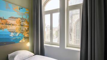 Habitación de hotel con una cama, un cuadro de un paisaje y vistas de edificios a través de la ventana.