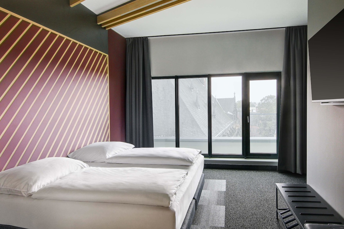 A modern hotel room with a large bed, a view of buildings through the window, and a unique striped wall design.