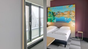 Habitación de hotel con cama, ventana que ofrece vistas a edificios y un cuadro colorido en la pared.