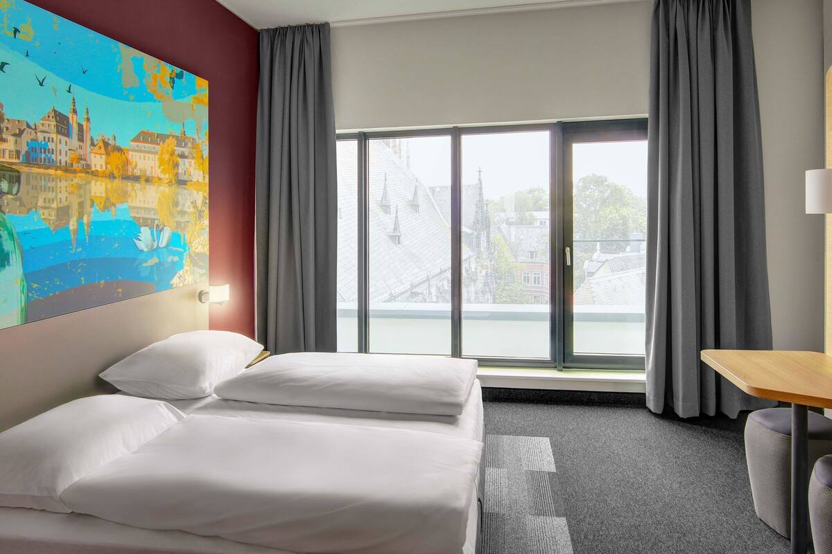 A hotel room with a bed, a desk, and a large window with curtains.