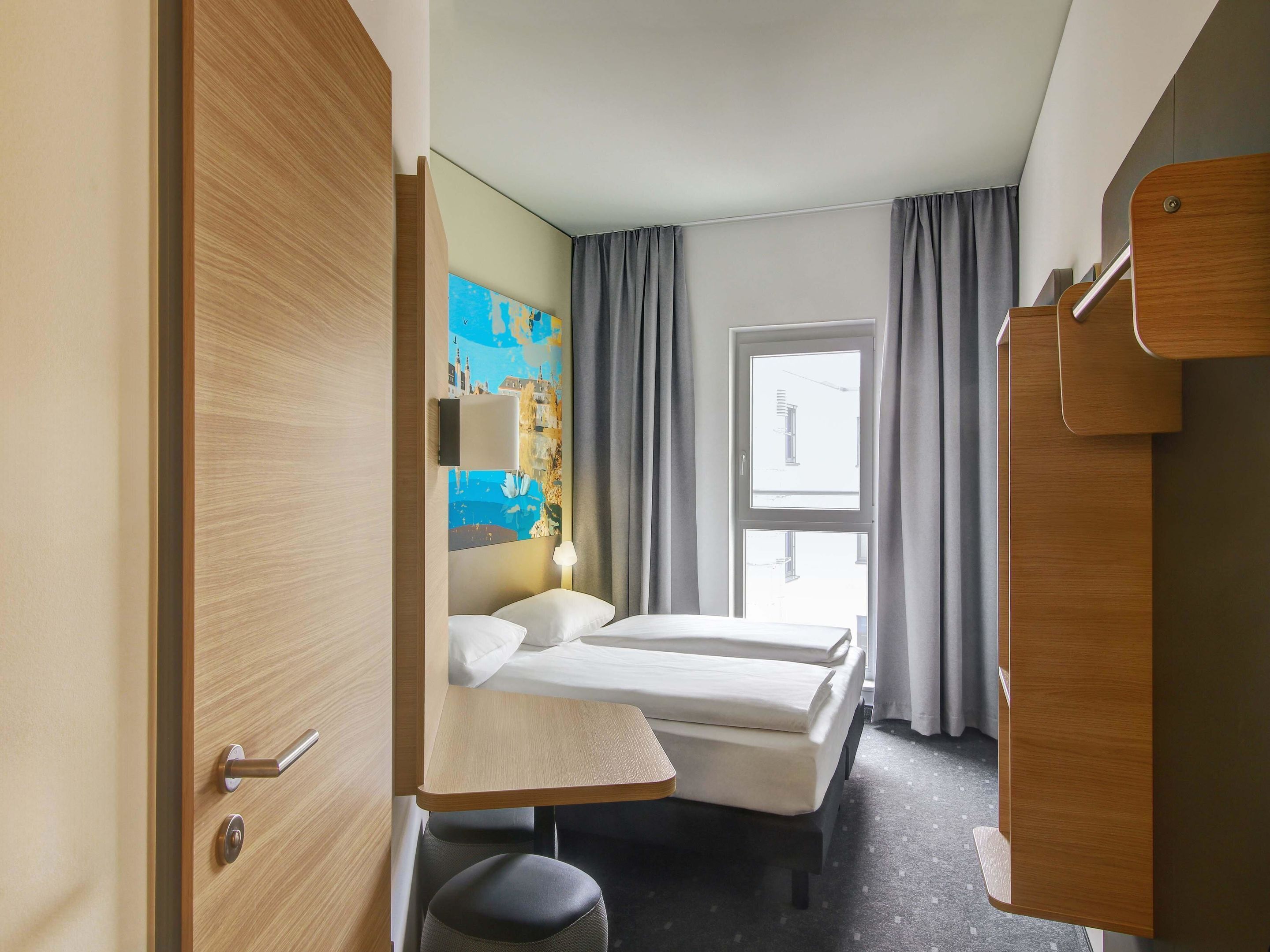 Habitación de hotel con dos camas, un escritorio, una silla, una ventana con cortinas y un cuadro en la pared.