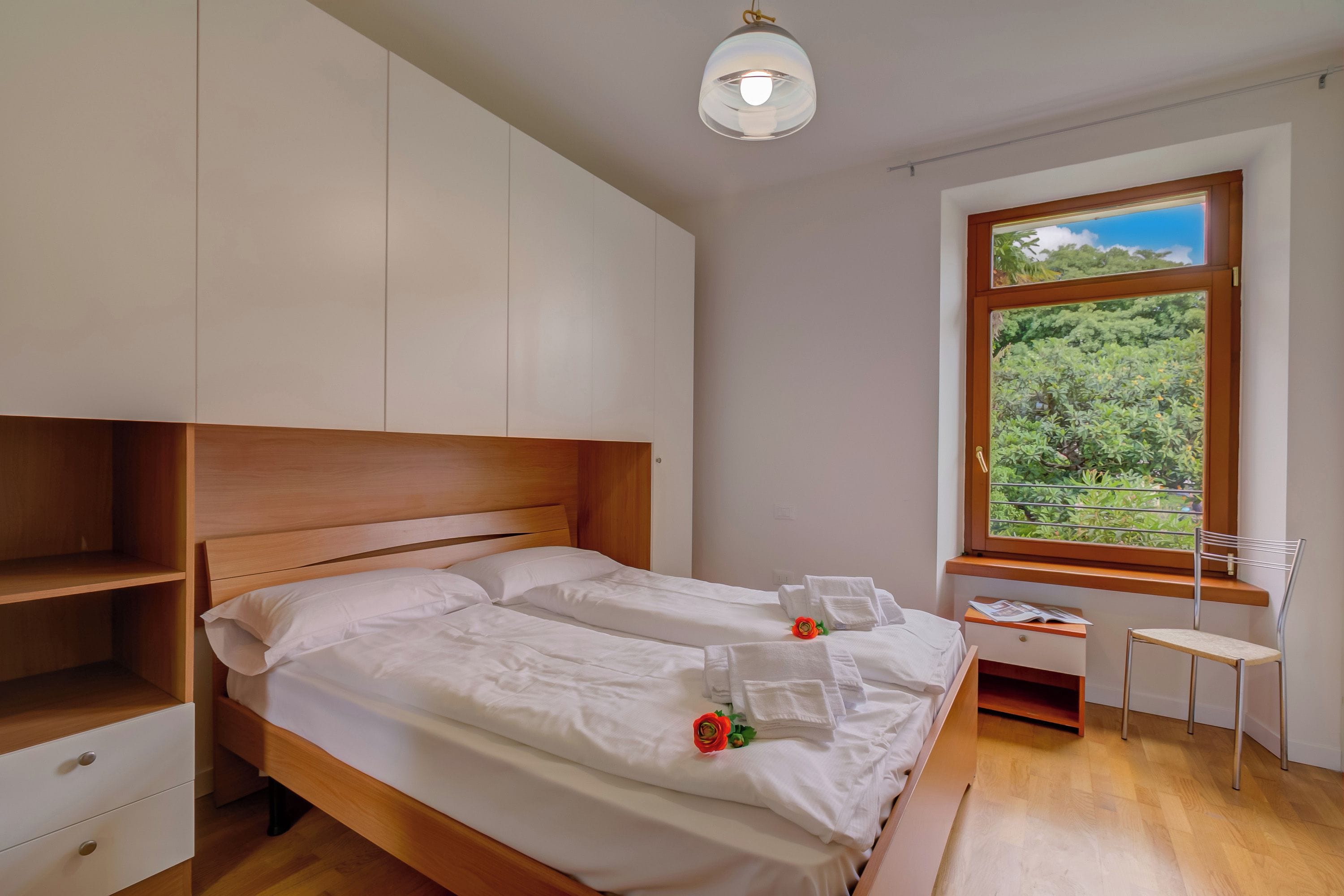A bedroom with a wooden bed, white bedding, a nightstand, a chair, a window with a view of greenery, and a ceiling light.