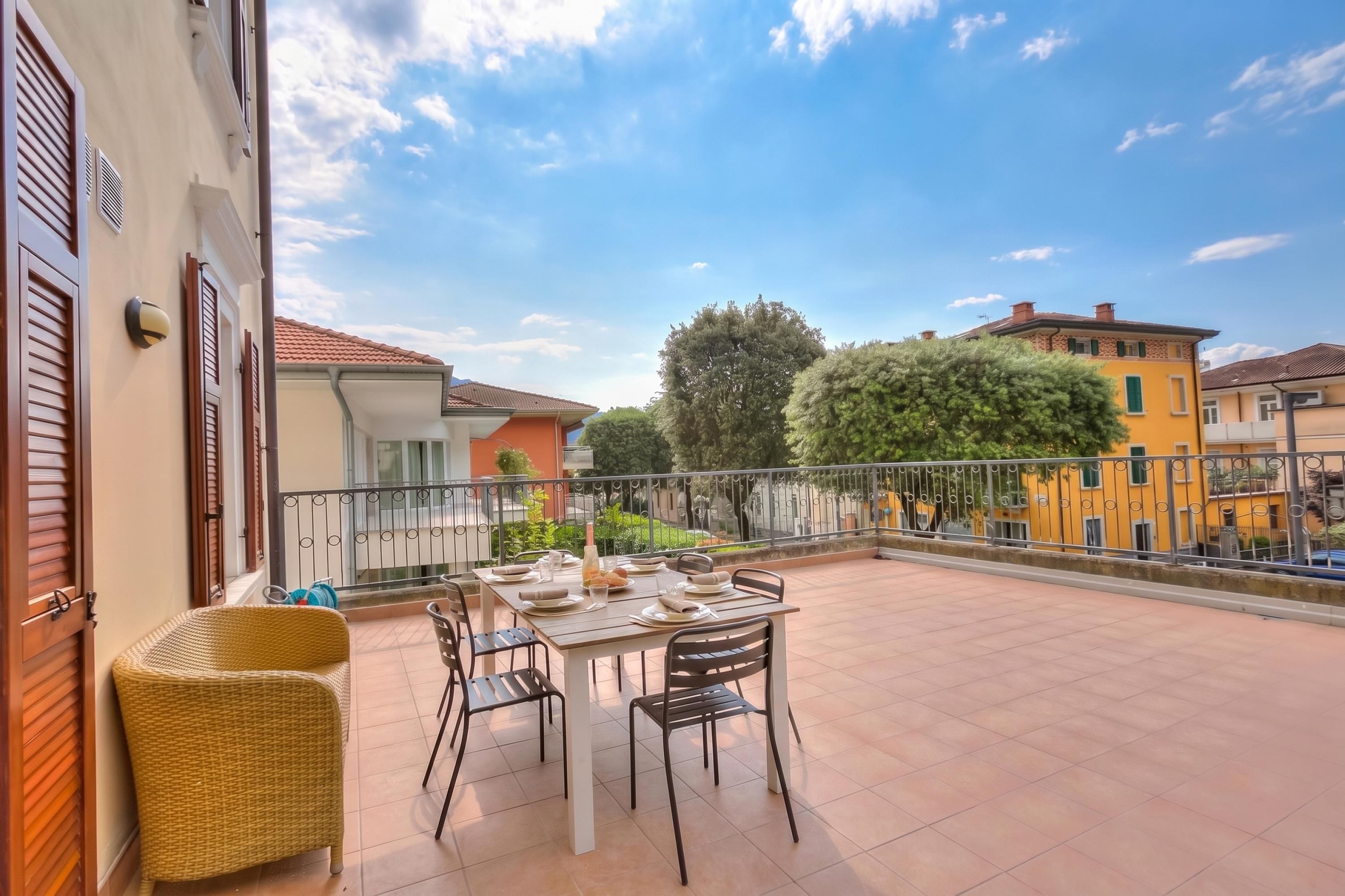 A balcony with a table set for dining, surrounded by wicker furniture, and overlooking a residential area with trees and buildings.
